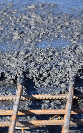 Das Bild zeigt grobkörnigen, dunkelgrauen Schwerbeton, der frisch auf eine Baustelle gegossen wird. Im Vordergrund liegen rostfarbene Stahlbewehrungen, die teilweise bereits vom Beton umschlossen sind. Die Betonmasse wirkt sehr dicht und schwer, einzelne große Zuschlagstoffe sind deutlich zu erkennen. Schwerbeton wird durch den Einsatz von besonders schweren Gesteinskörnungen hergestellt und kommt vor allem dort zum Einsatz, wo hohe Dichte und Strahlenschutz gefragt sind, etwa im Kraftwerks- oder Strahlenschutzbau.

                                    